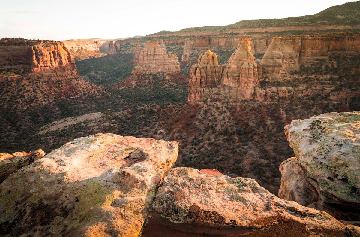 Colorado National Monument featuring towering red rock formations, rugged cliffs, and desert vegetation. The landscape highlights unique geological structures, layered rock textures, and panoramic views under a clear sky